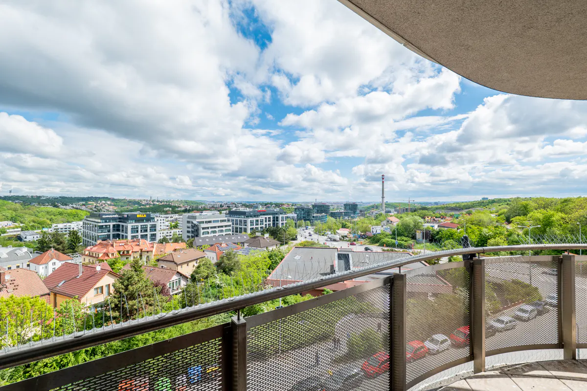 View from a balcony with a metal railing overlooking a city with buildings, trees, and a cloudy blue sky.