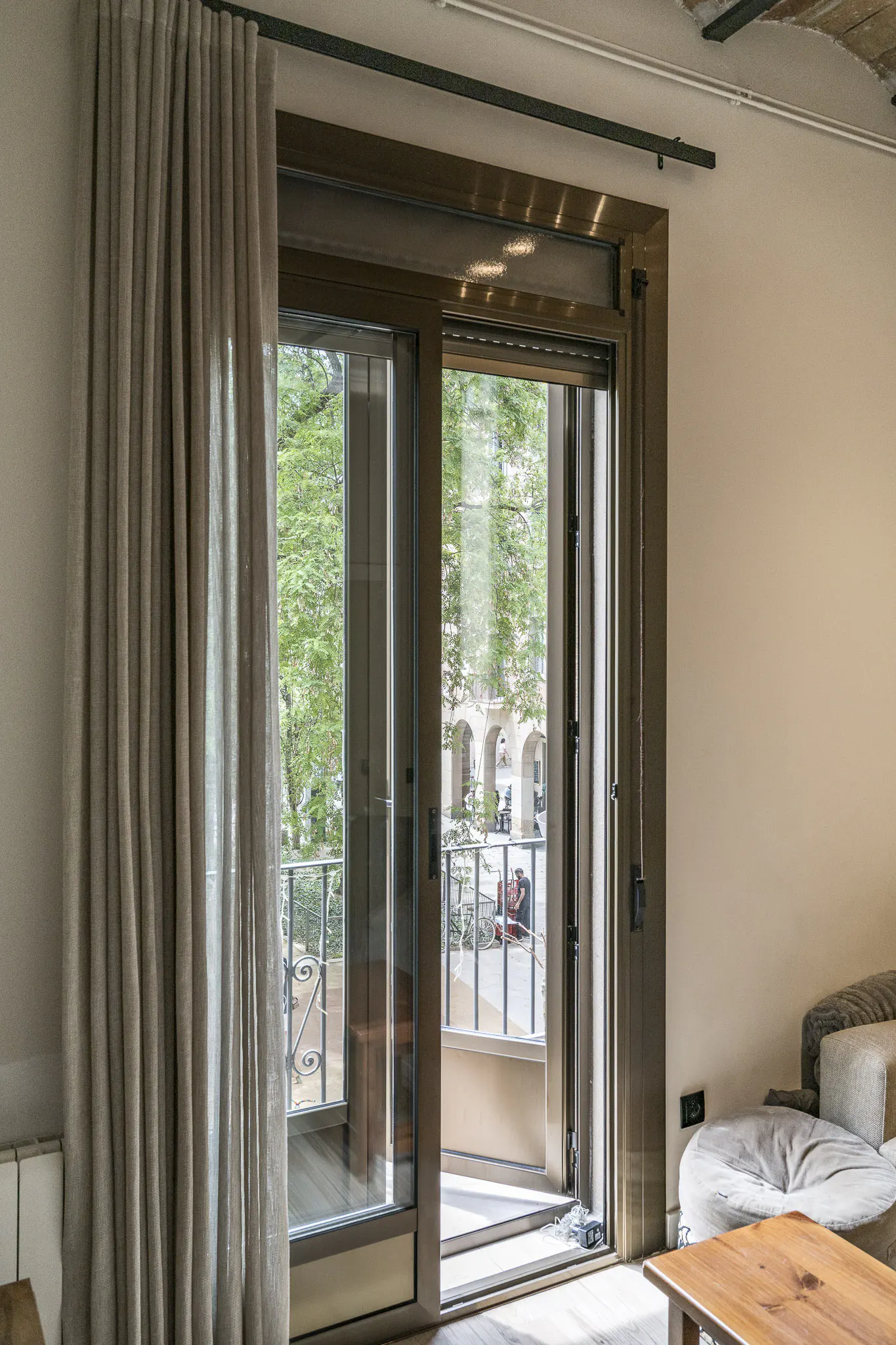 Sliding glass door with beige curtains opening to a balcony overlooking a tree-lined street.