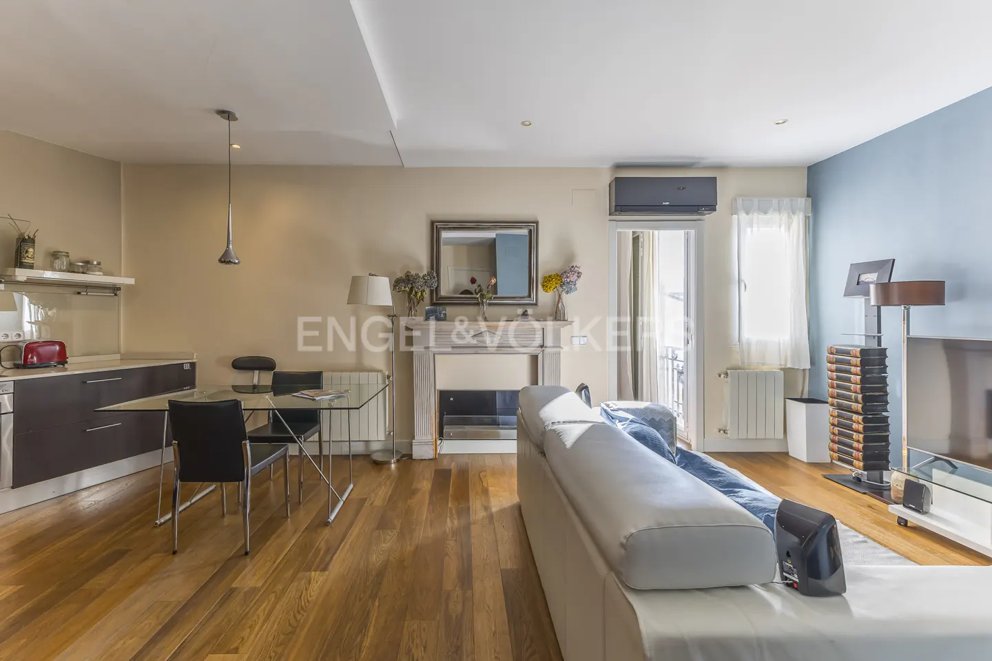 A wide shot of a modern apartment with a kitchen, dining area, and living room. Hardwood floors, white sofa, and a fireplace are visible.