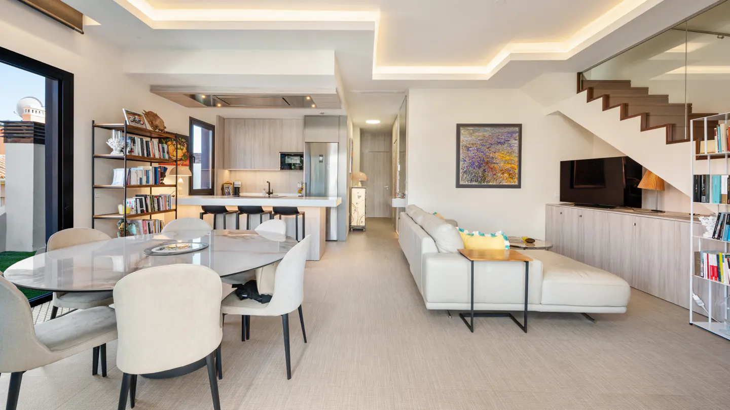 Modern open-plan living area with a dining table, white sofa, kitchen island, and a staircase leading to an upper level.