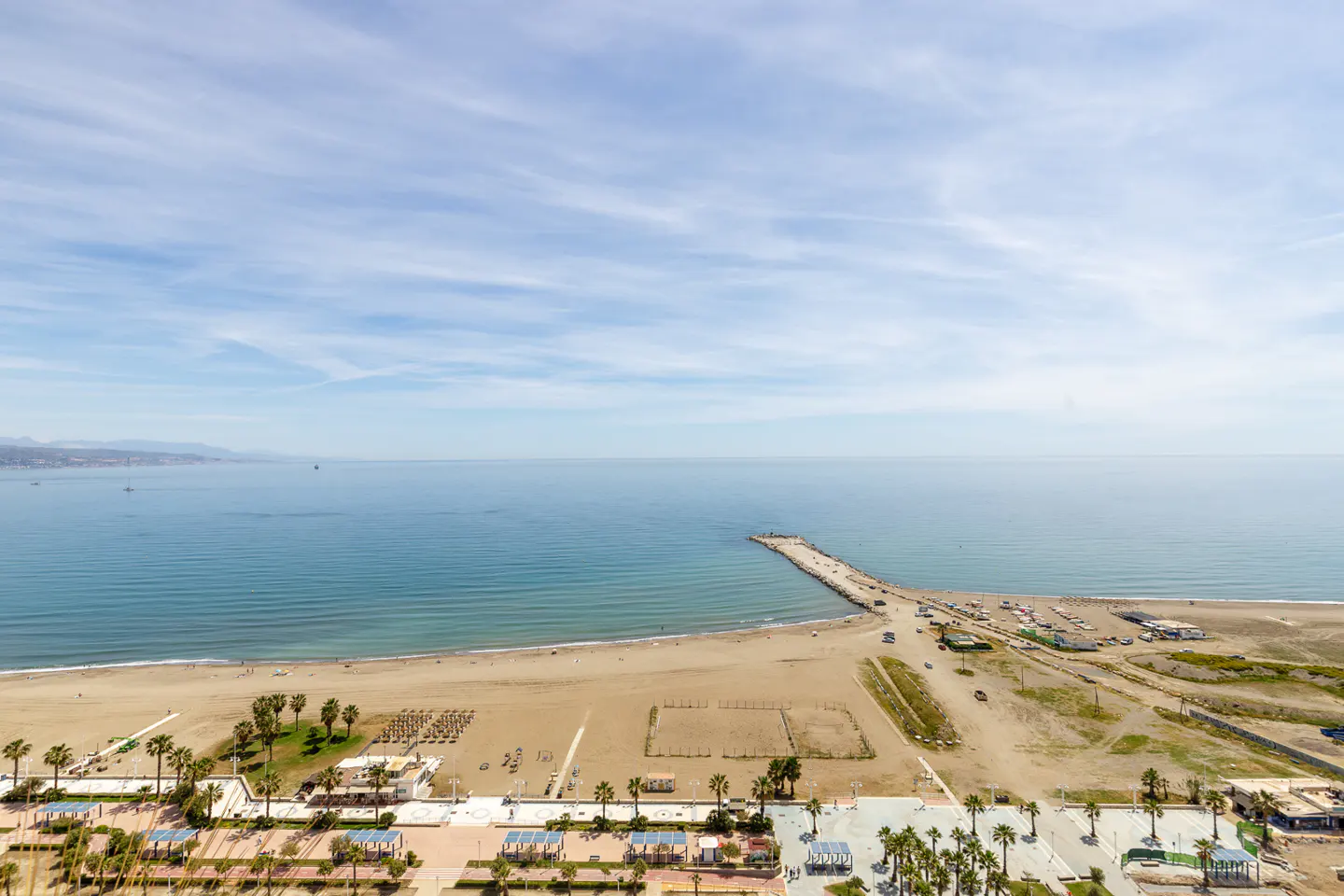 Aerial view of a sandy beach with palm trees, a pier extending into the blue sea, and a partly cloudy sky.