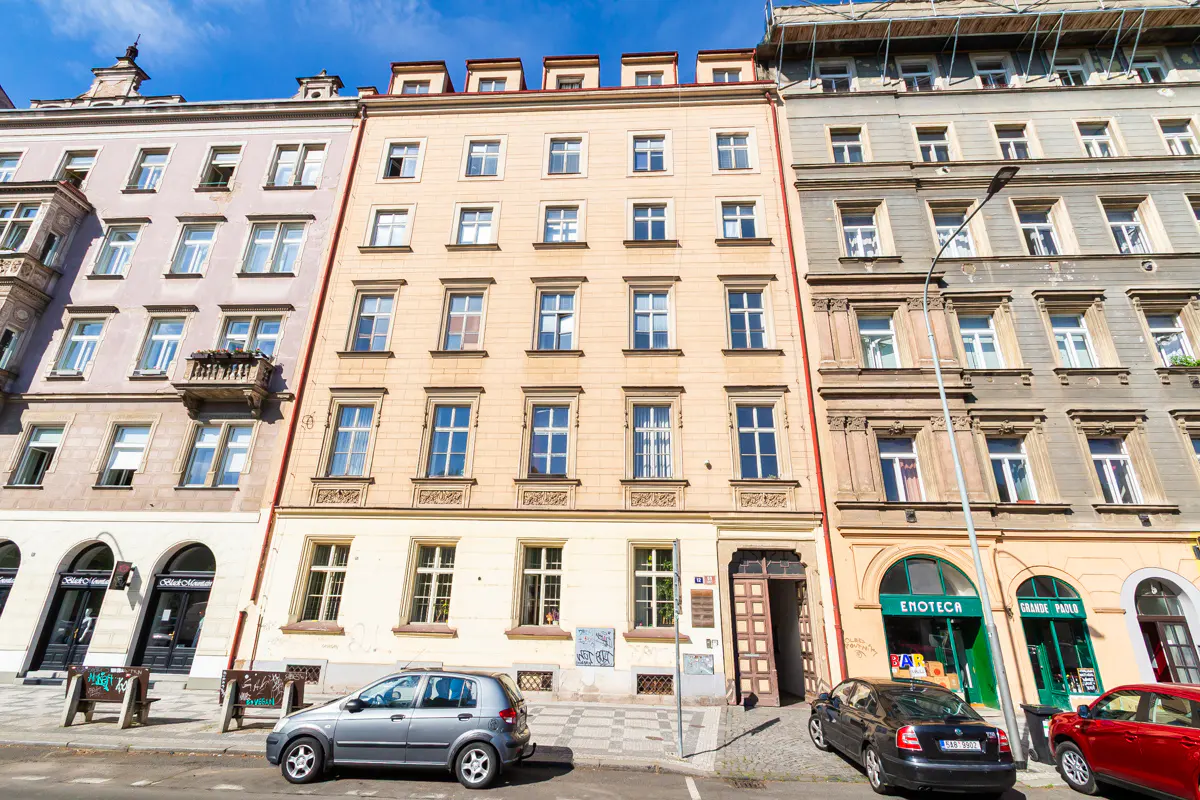 Street view of a tall, narrow, light-yellow building with many windows, flanked by similar buildings and parked cars.