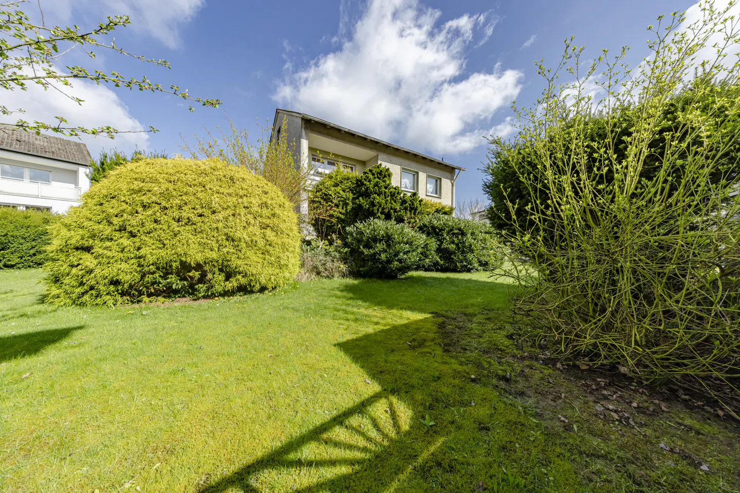 A house with a green lawn and bushes under a blue sky with white clouds.