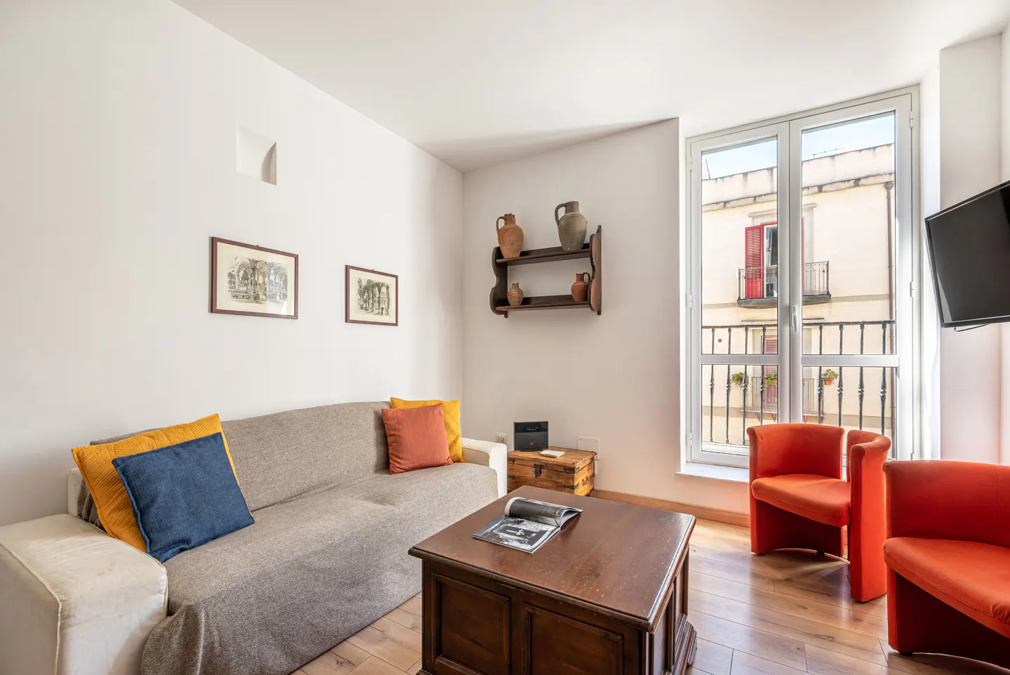 Living room with a beige sofa, orange chairs, and a wooden coffee table. A window shows a building with a red door.