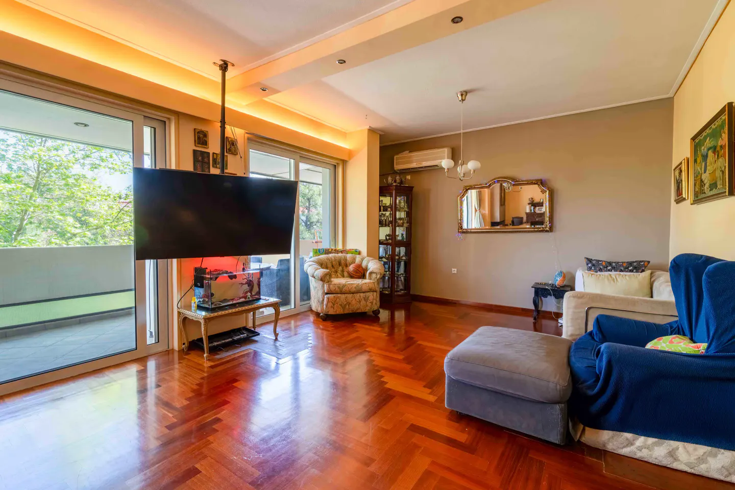 A living room with hardwood floors, a large TV, and a balcony with a view of trees.