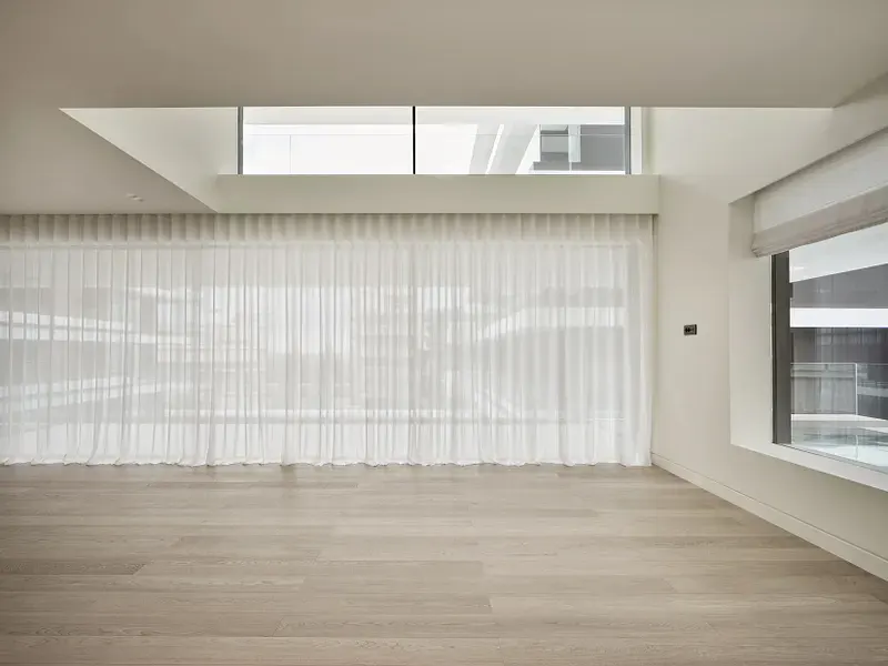 Bright, empty room with light wood floors, white walls, and sheer white curtains covering a large window. A smaller window is on the right.