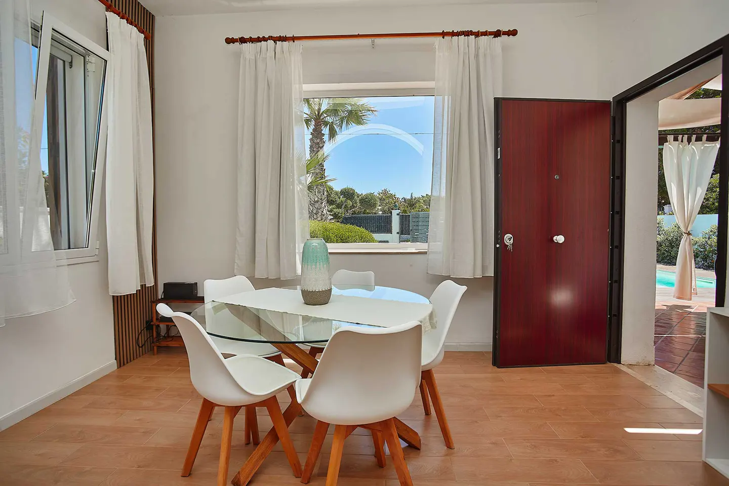 Bright dining room with a glass table, white chairs, and a view of a pool through an open door. White curtains frame a window with a palm tree view.