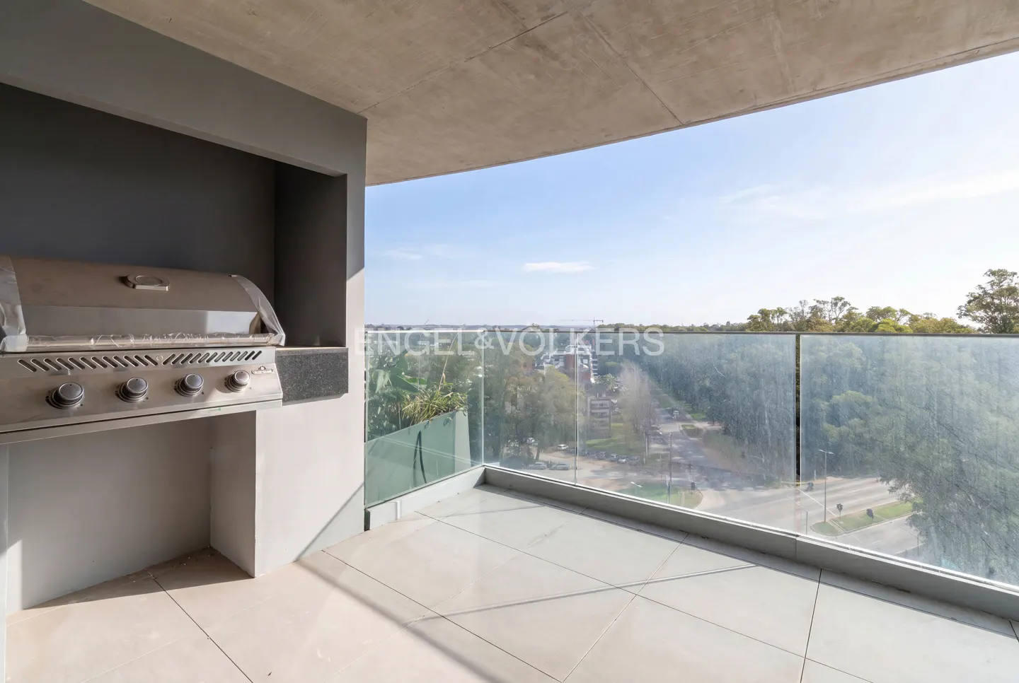 Modern balcony with a built-in stainless steel grill, light tile flooring, and a glass railing overlooking a tree-lined road.