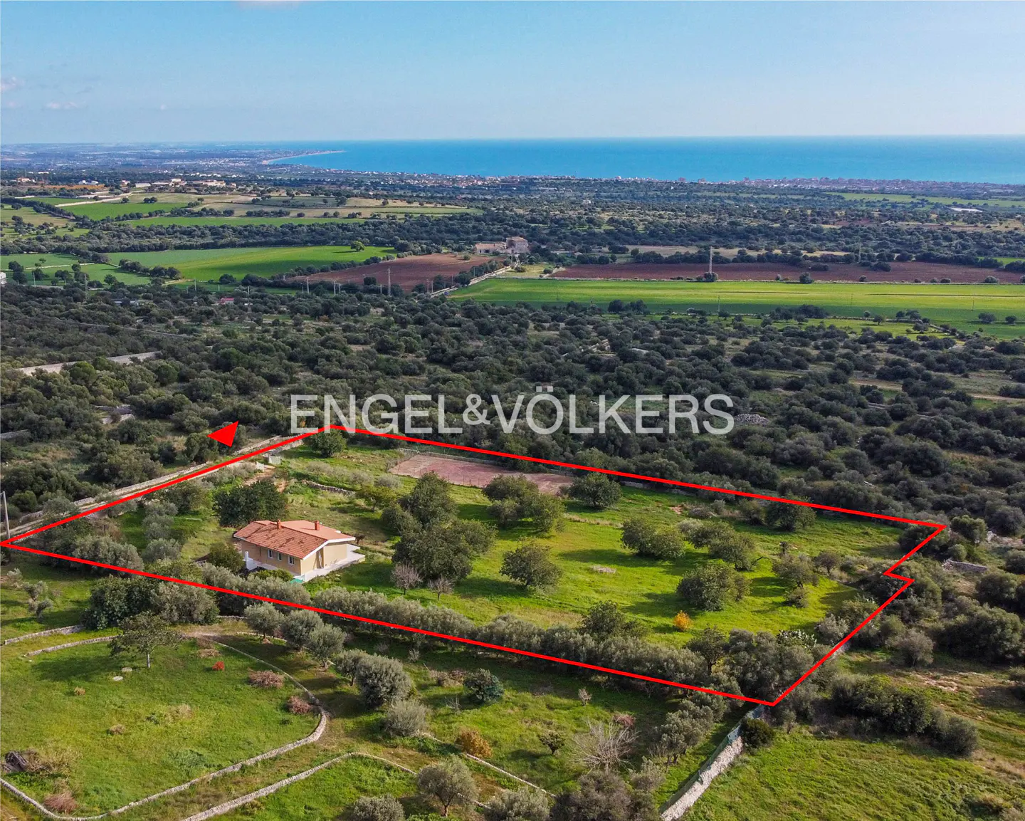 Aerial view of a property outlined in red, featuring a house, trees, and the Engel & Völkers logo, with the sea in the background.