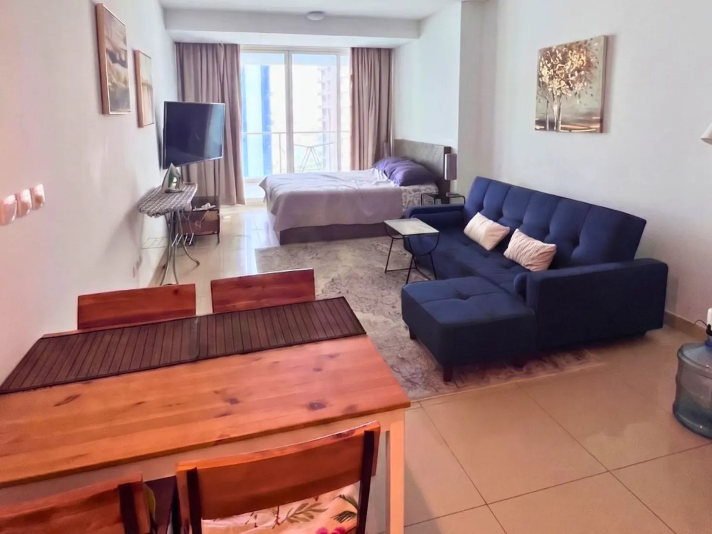 Studio apartment with a wooden dining table, blue sofa, bed, TV, and balcony with curtains.