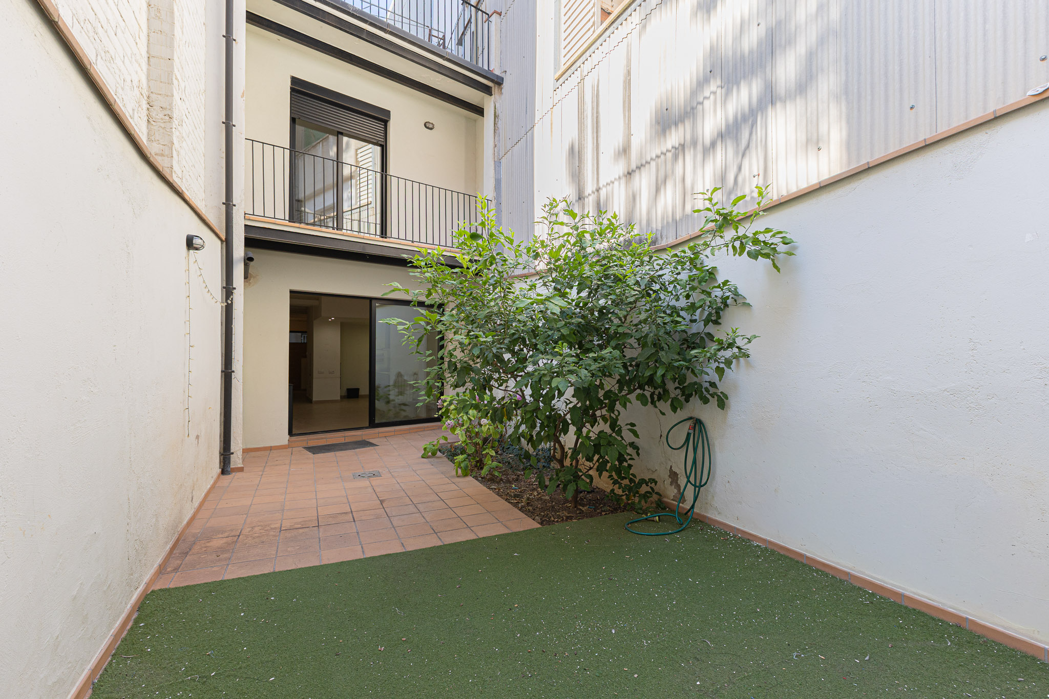 A small urban patio with tiled flooring, a patch of artificial grass, a green shrub, and a garden hose against white walls.