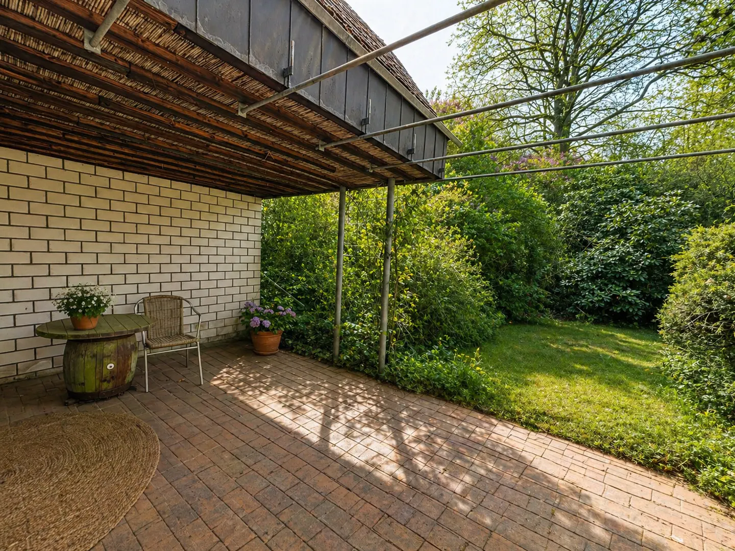 Covered patio with brick floor, white brick wall, and lush green garden. A barrel table, chair, and potted flowers add charm.