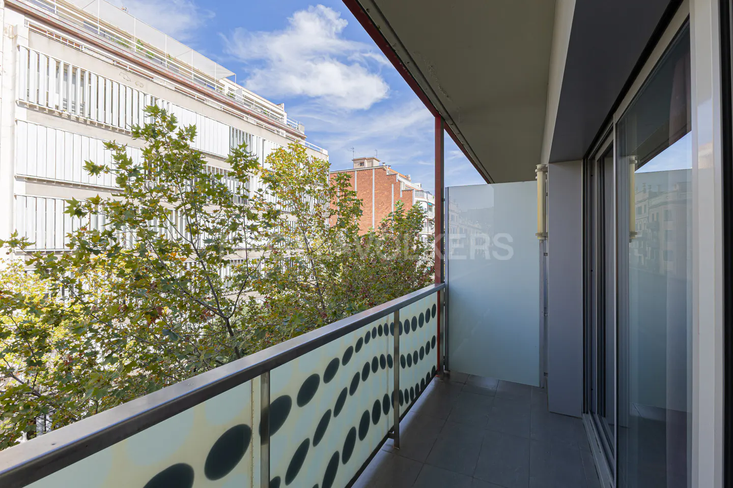 Balcony view with patterned glass railing, trees, and buildings under a blue sky. Sliding glass doors lead inside.