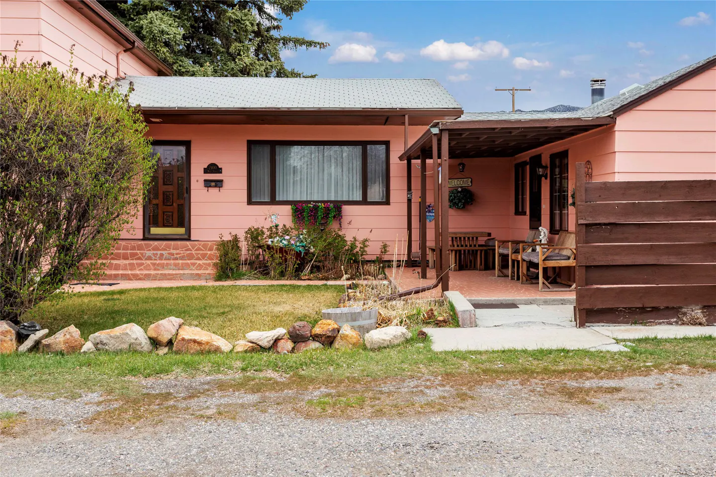 Single-story pink house with a covered patio, wooden privacy fence, and a front yard with rocks and a small tree.