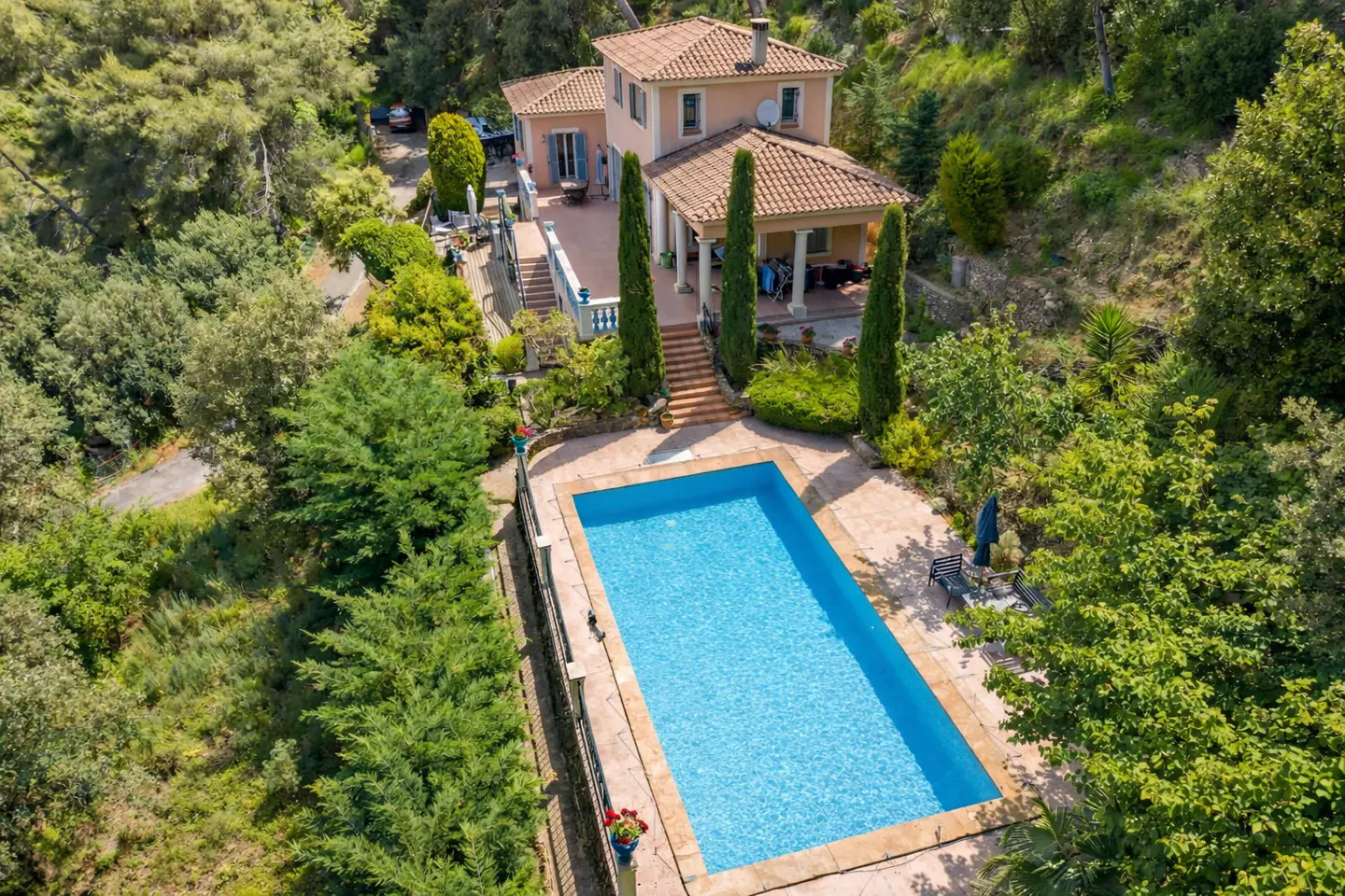 Aerial view of a peach-colored two-story house with a red tile roof and a blue rectangular swimming pool surrounded by trees.