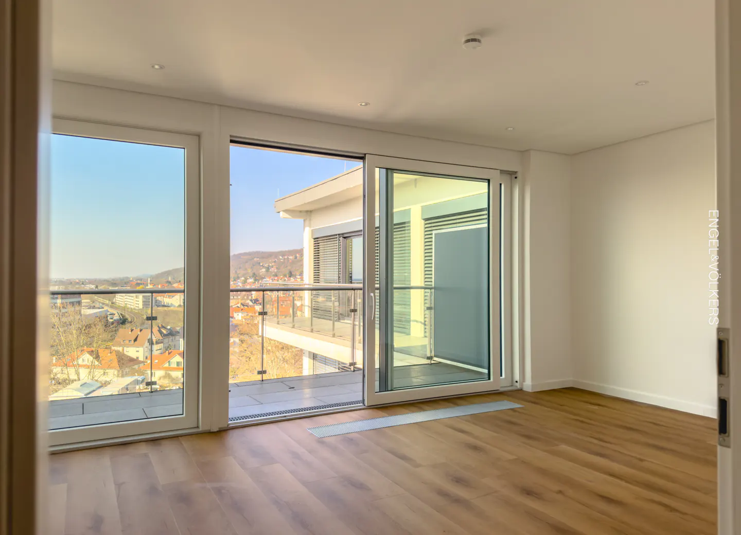 Bright, empty room with wood floors and white walls. Sliding glass doors open to a balcony with a city view.