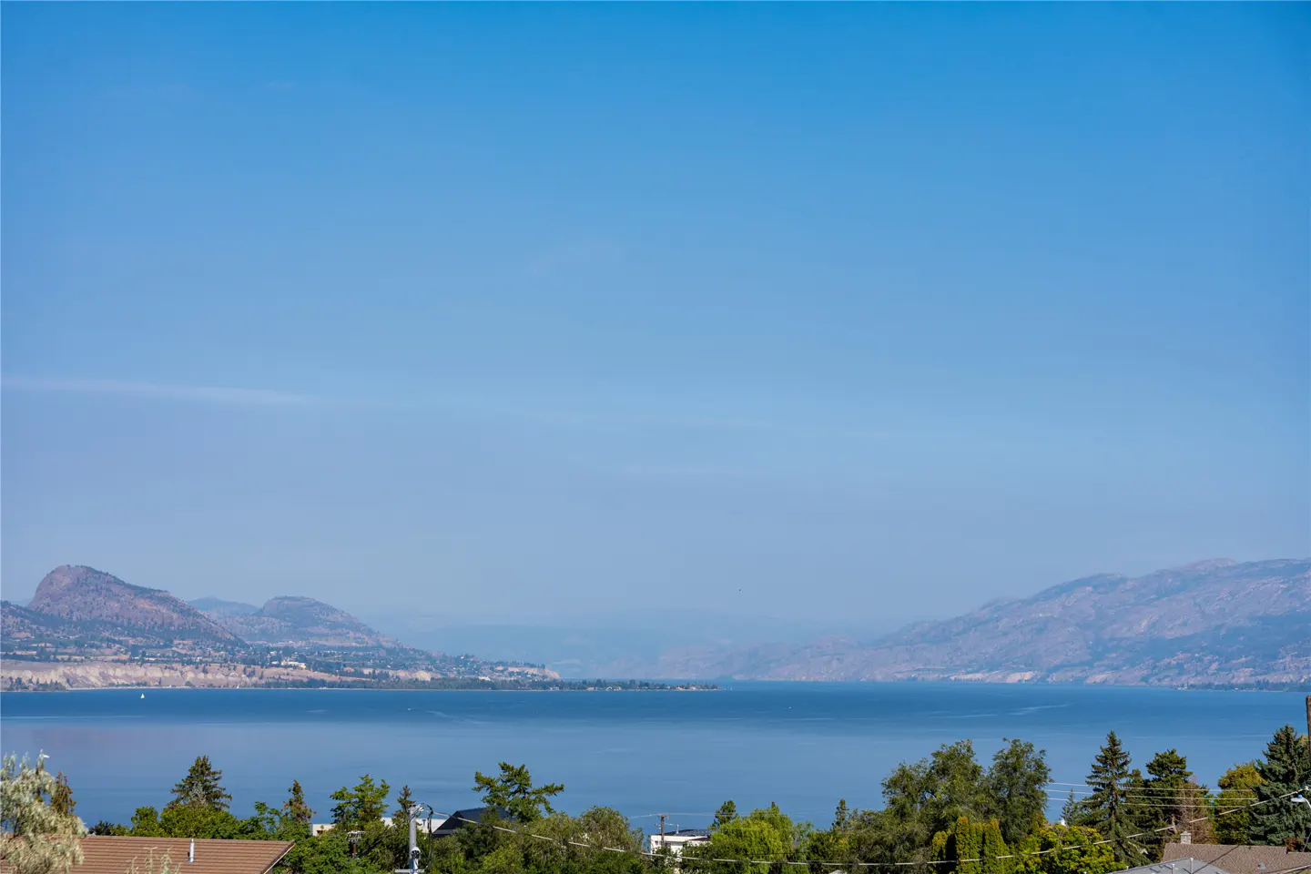 Scenic view of a lake surrounded by mountains under a clear blue sky, with trees and rooftops in the foreground.