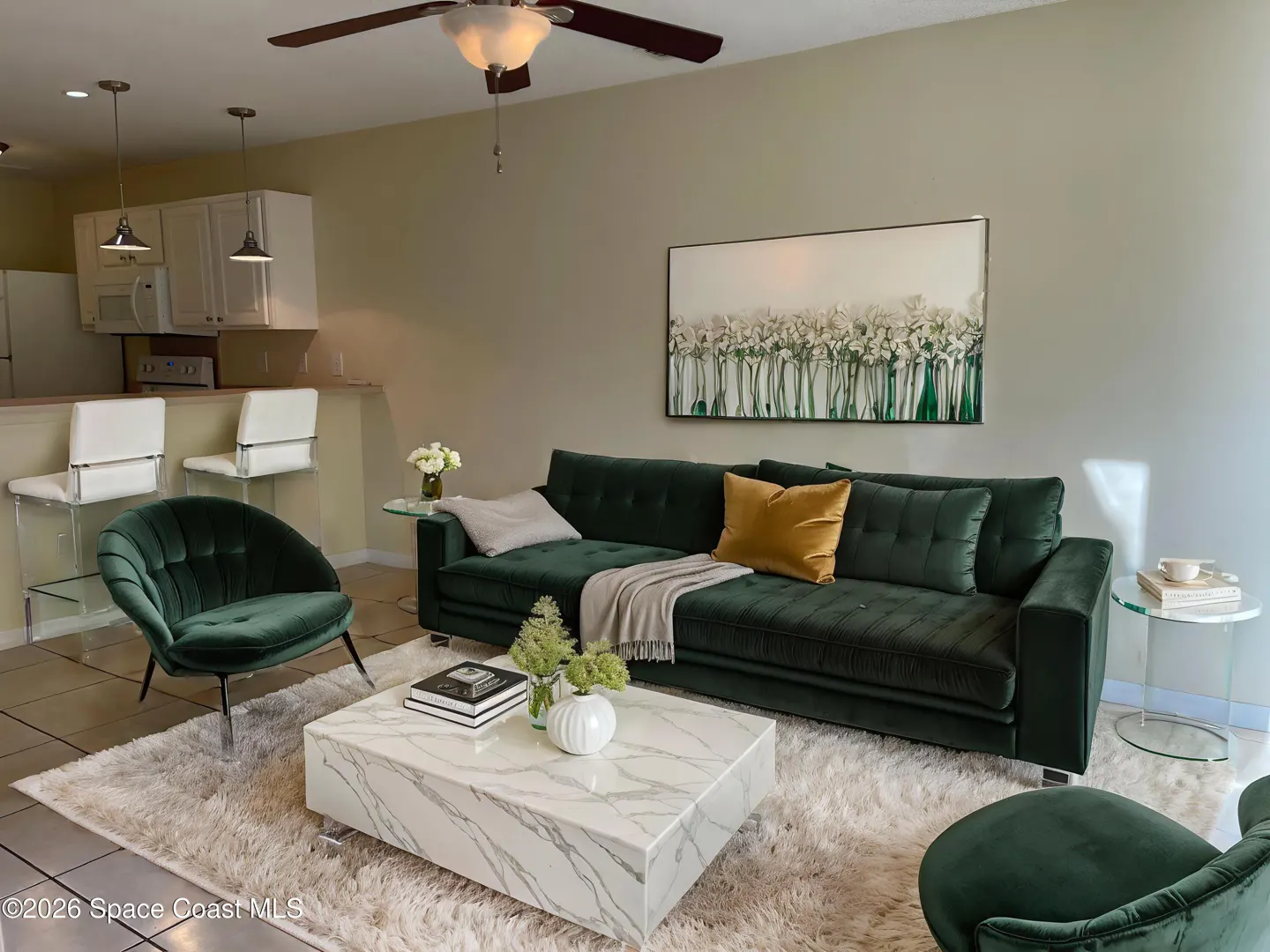 Living room with green velvet sofa, chair, and marble coffee table on a white shag rug. Kitchen with white cabinets and bar stools in the background.