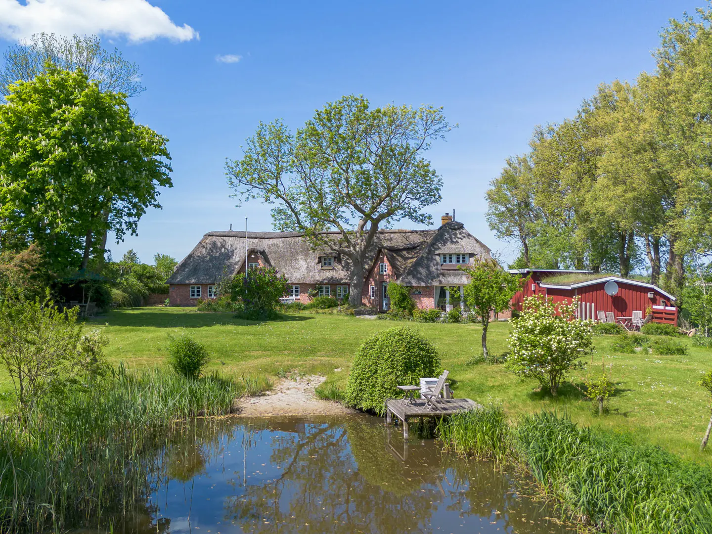 Idyllic country home with a thatched roof, surrounded by lush greenery and a serene pond under a clear blue sky.