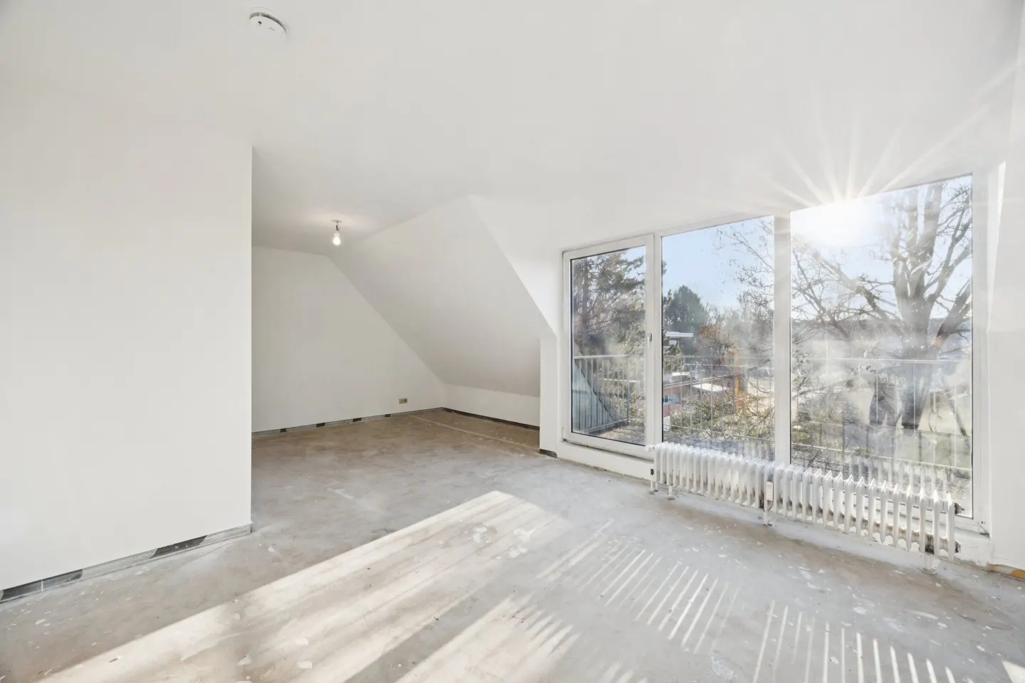 Bright, empty attic room with white walls, bare concrete floor, and large windows with a radiator. Sunlight streams in.