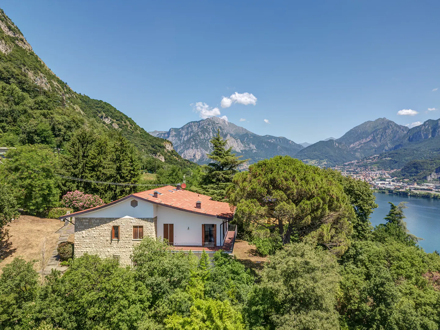 Scenic view of a stone house with a red tile roof, surrounded by lush green trees, mountains, and a blue lake under a clear sky.