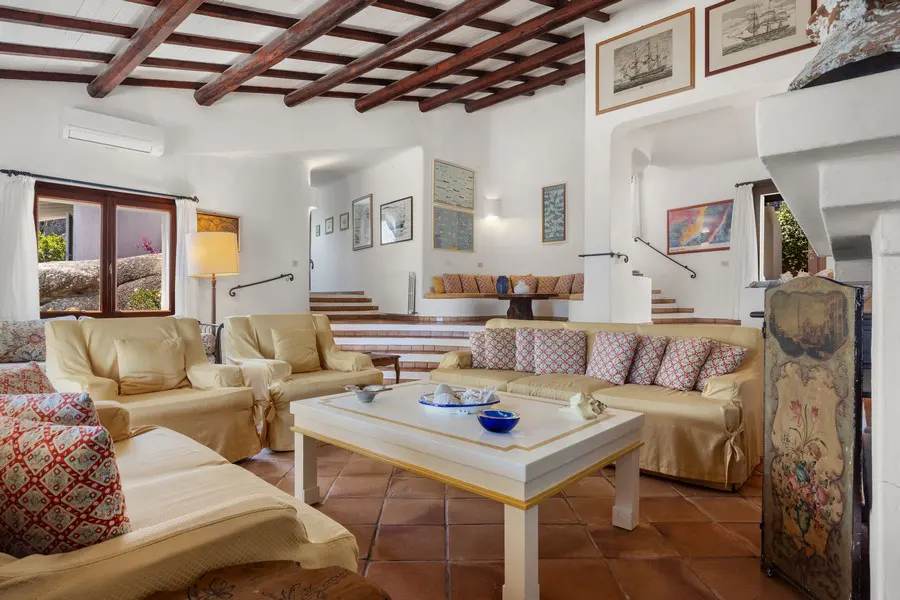 Bright living room with white walls, terracotta tile floor, and exposed wood beam ceiling. Cream-colored sofas and a white coffee table furnish the space.