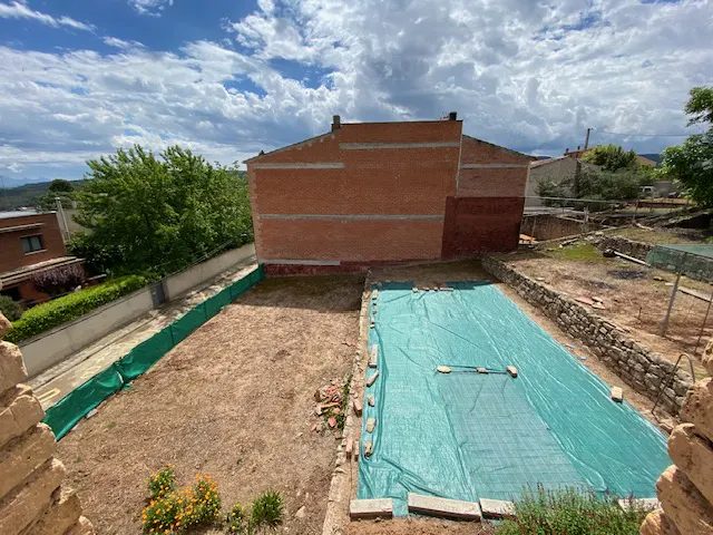 An aerial view of a backyard with a green tarp covering a pool, a brick building, and a stone wall.