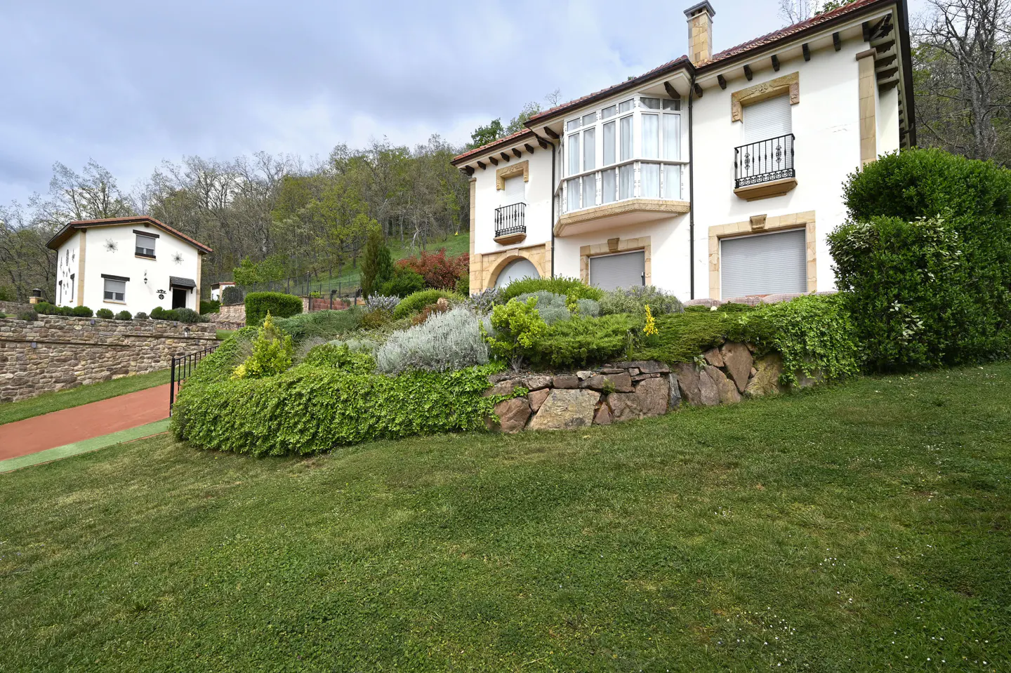 A two-story white house with a red tile roof sits on a green lawn with a stone retaining wall and lush landscaping.
