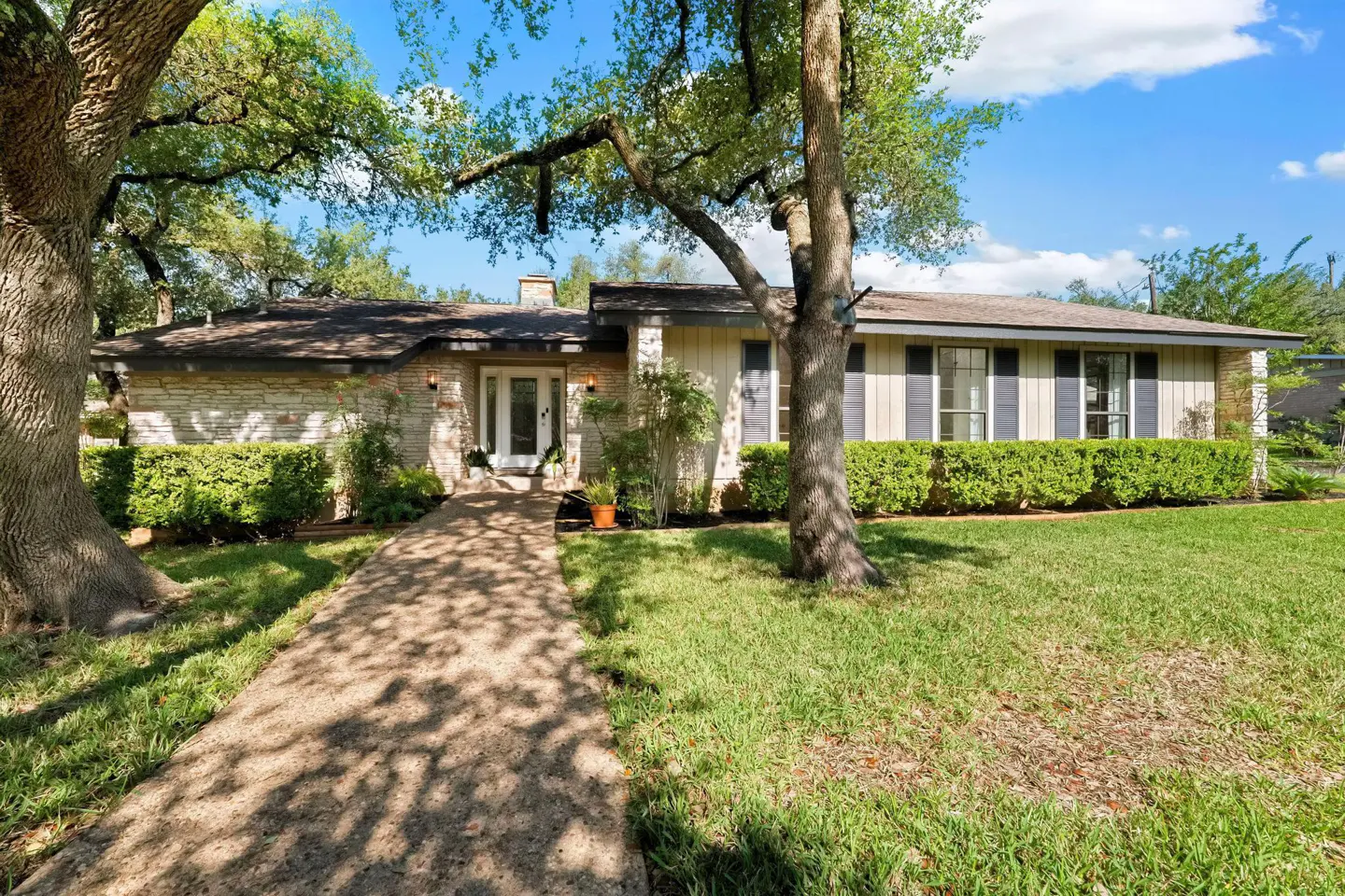A ranch-style house with a stone and beige facade, dark shutters, and a walkway leading to the front door. Mature trees frame the house.