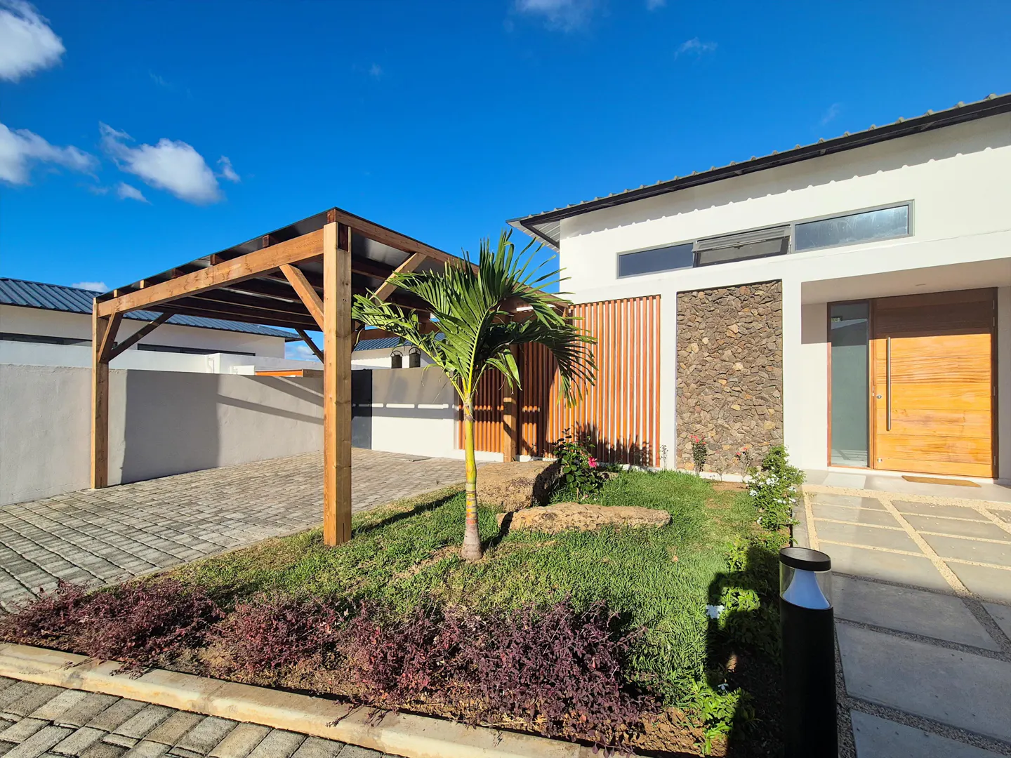 Modern home exterior with a wooden carport, stone accents, and a small palm tree in the front yard under a blue sky.