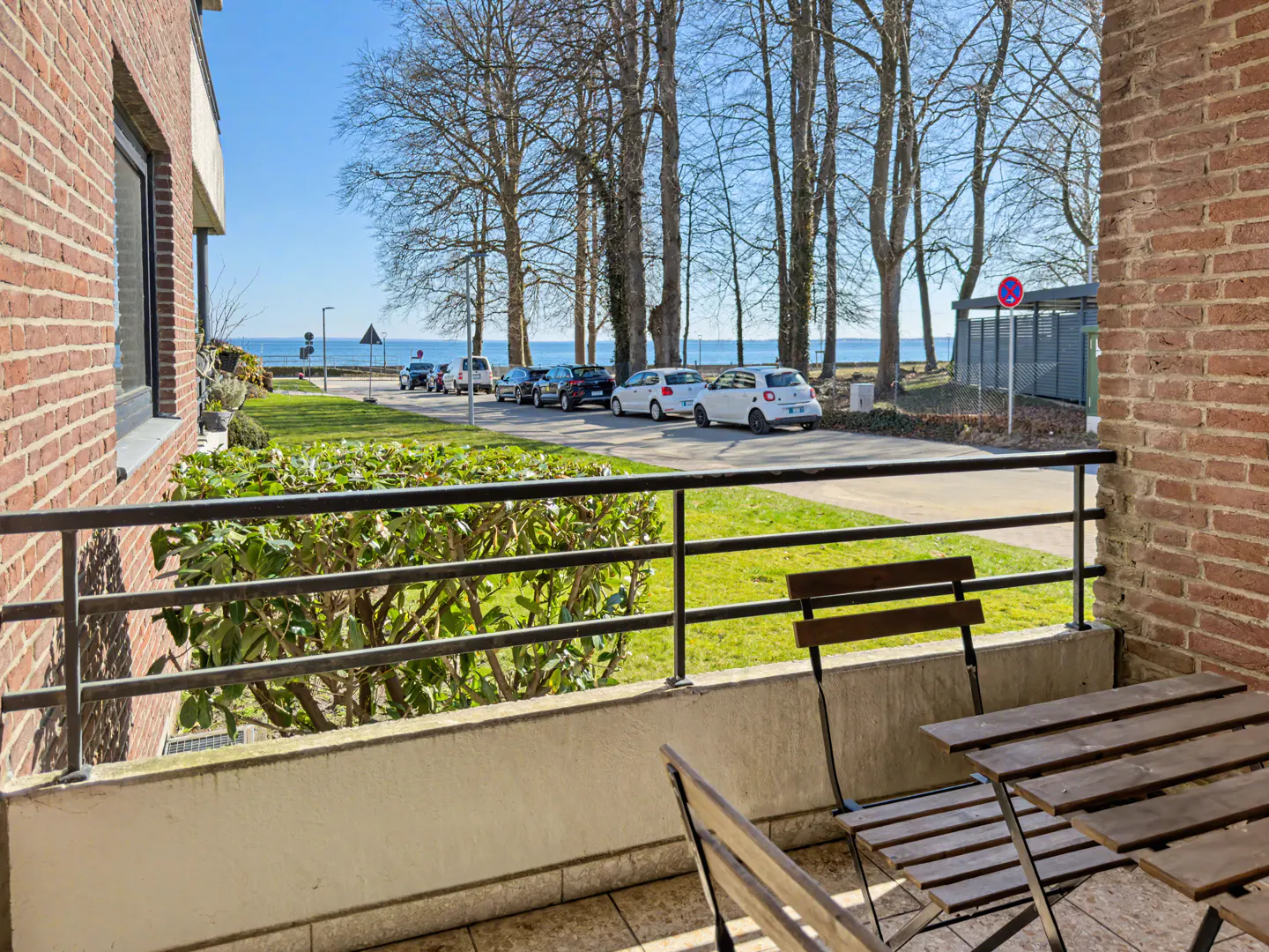 Balcony view with a wooden table and chairs, overlooking a street with parked cars, trees, and the ocean in the background on a sunny day.