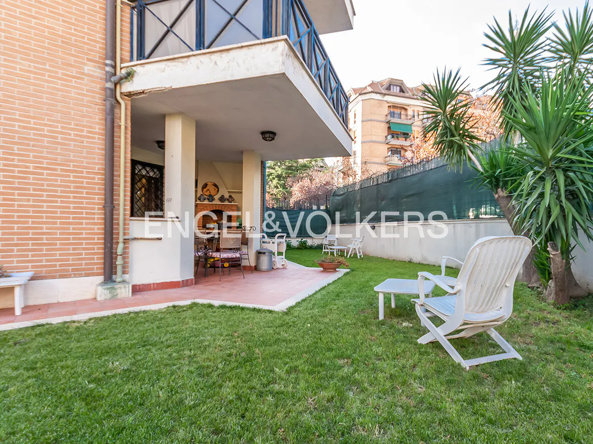 Backyard of a brick house with a covered patio, grill, lawn, and white plastic chairs. Engel & Volkers logo is visible.