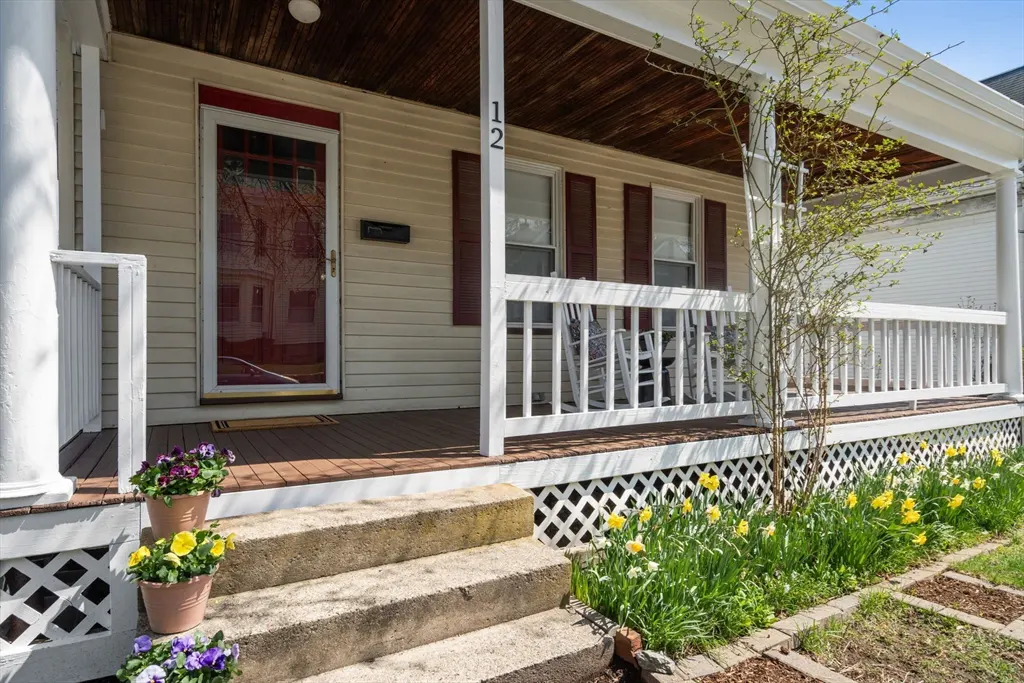 Exterior of a light yellow house with a red door and shutters, a porch with white railings, and flower pots on the steps.