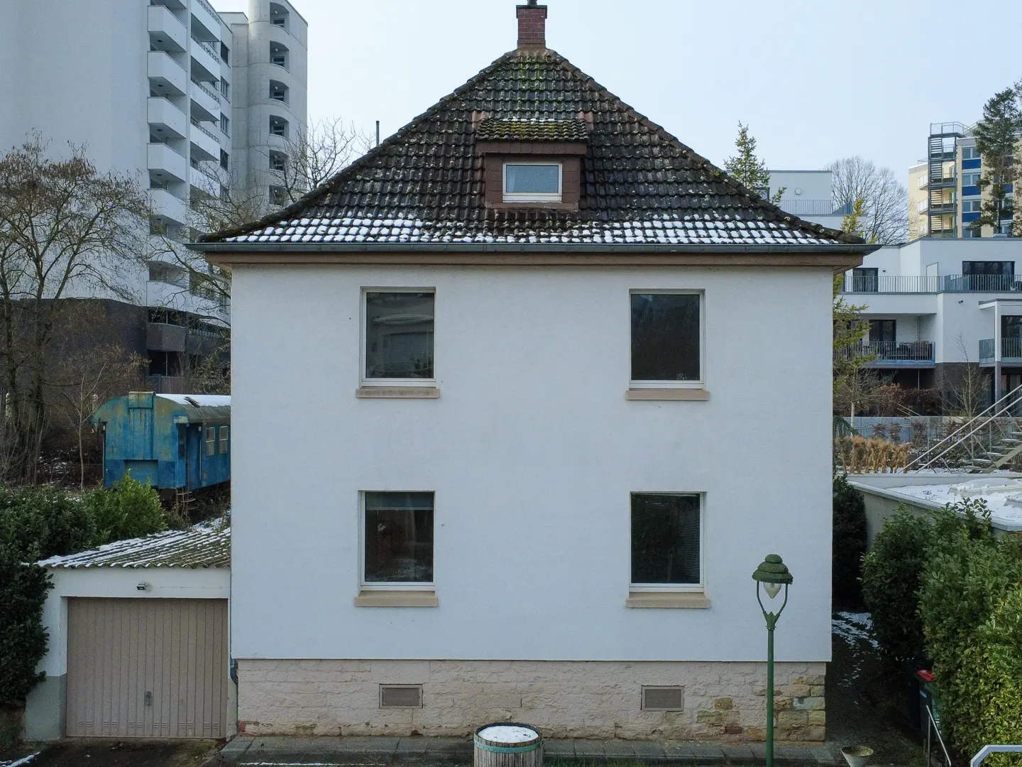 Two-story white house with a brown tiled roof and four windows. A garage is on the left, and a blue train car is in the background.