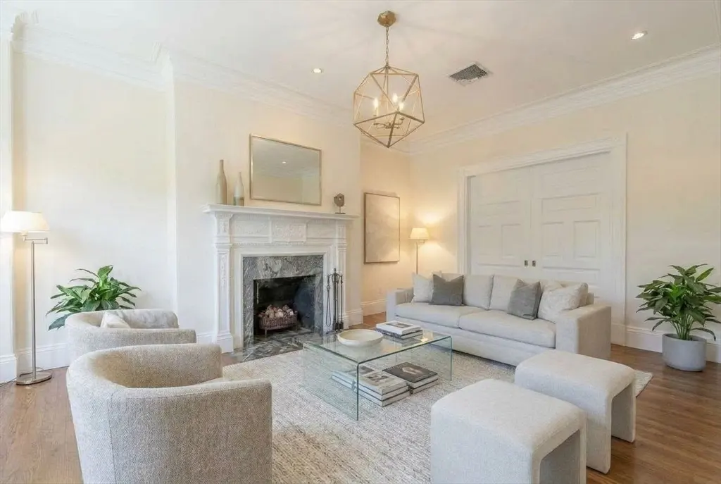Bright living room with a marble fireplace, white sofa, two chairs, and a glass coffee table. A gold geometric chandelier hangs above.