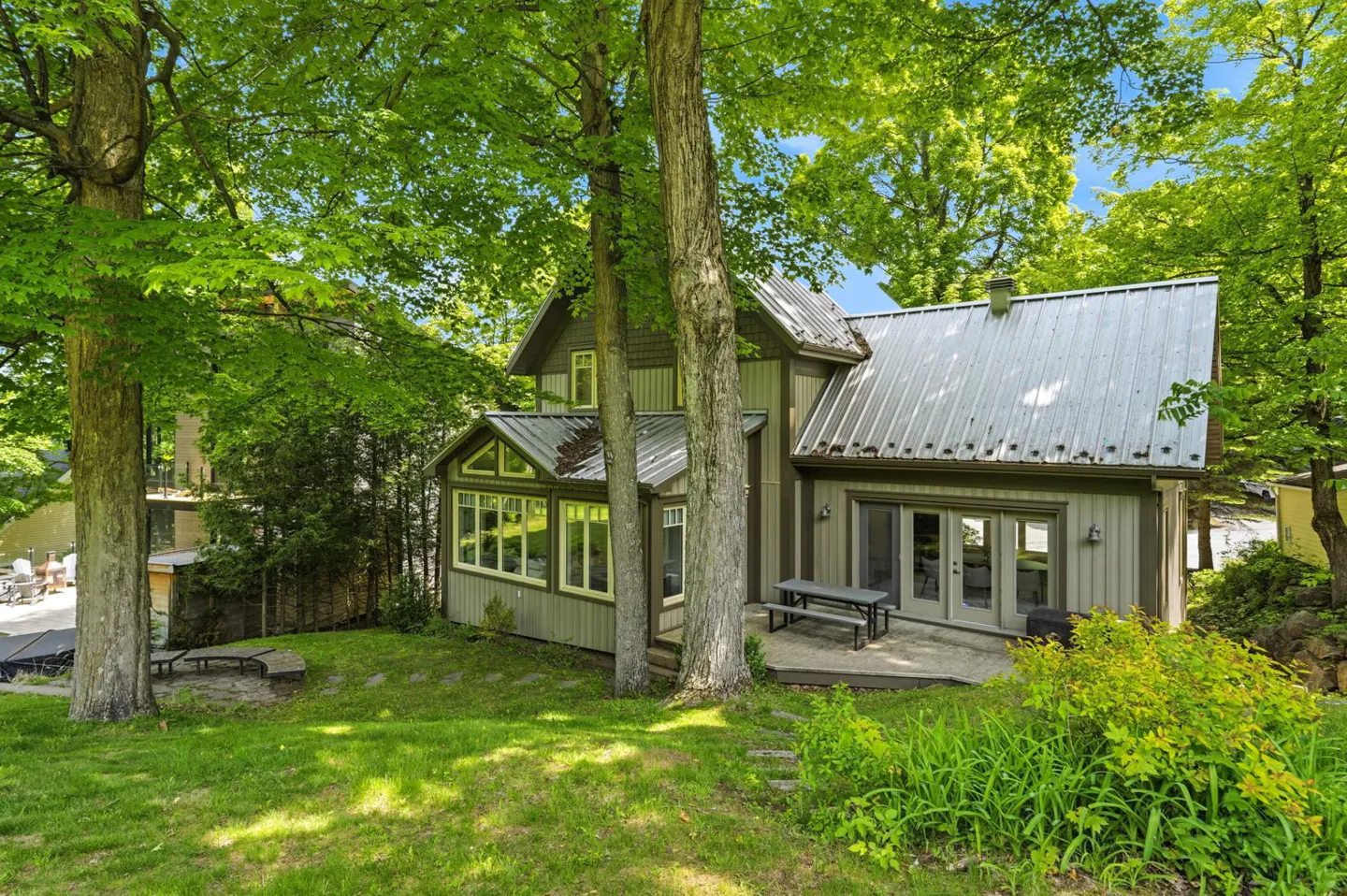 A two-story home with a metal roof and green siding, nestled in a lush, tree-filled yard with a patio and picnic table.