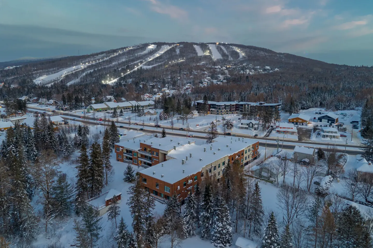 Aerial view of a brick apartment building covered in snow, with a ski hill in the background.