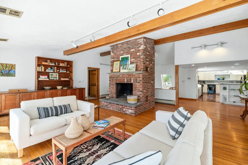 Bright living room with hardwood floors, white sofas, and a brick fireplace. A wooden bookshelf and beams add warmth to the space.