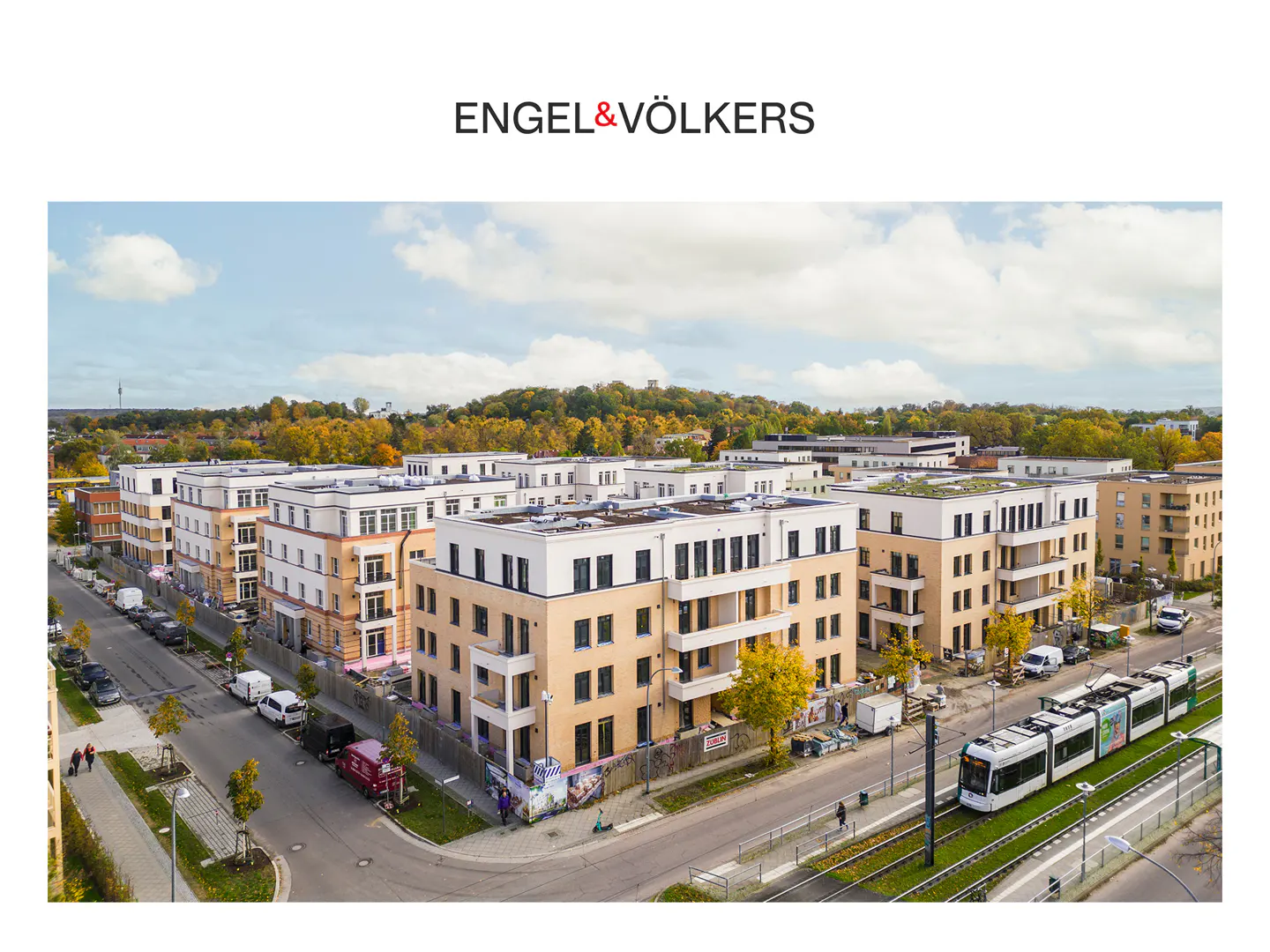 Aerial view of modern apartment buildings with beige walls and white roofs, a tram on the right, and trees in the background.