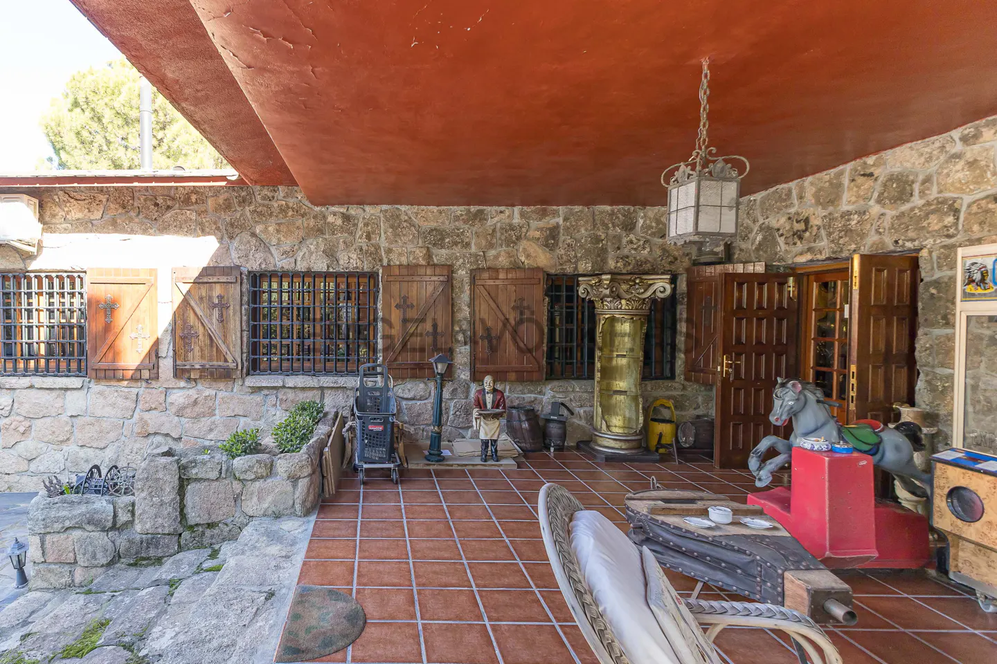 Covered patio with stone walls, red ceiling, and terracotta tile floor. Windows have iron bars and wooden shutters. Furnishings and decor are eclectic.