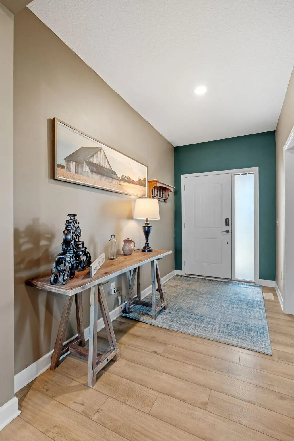 Entryway with wood floors, a blue rug, and a white front door. A wooden table with a lamp and decor sits against a tan wall with a barn picture.