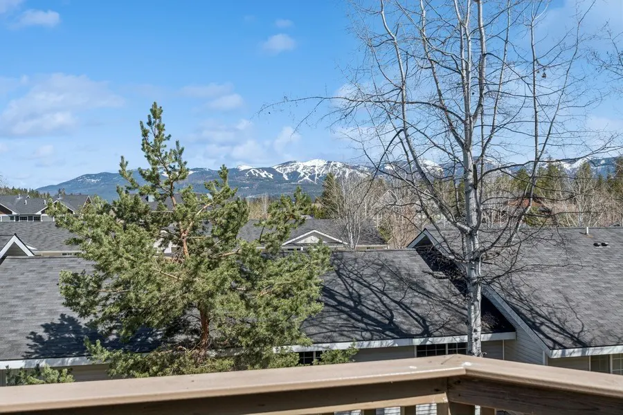 View from a deck overlooking rooftops, trees, and snow-capped mountains under a blue sky.