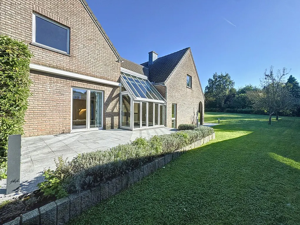 Brick house with a glass sunroom extension, a stone patio, and a large green lawn under a blue sky.