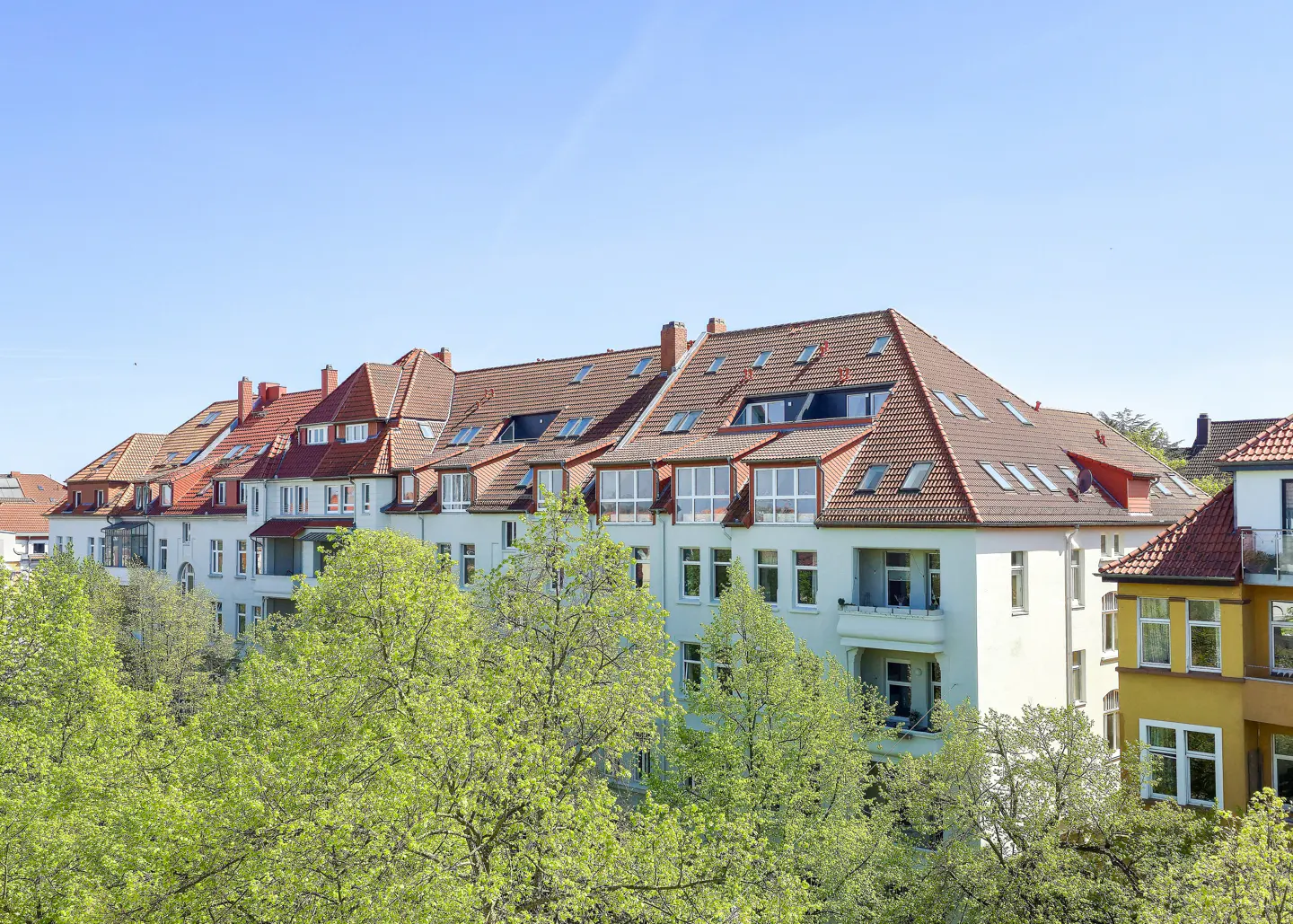 Row of multi-story apartments with red tile roofs and white facades, partially obscured by green trees against a blue sky.