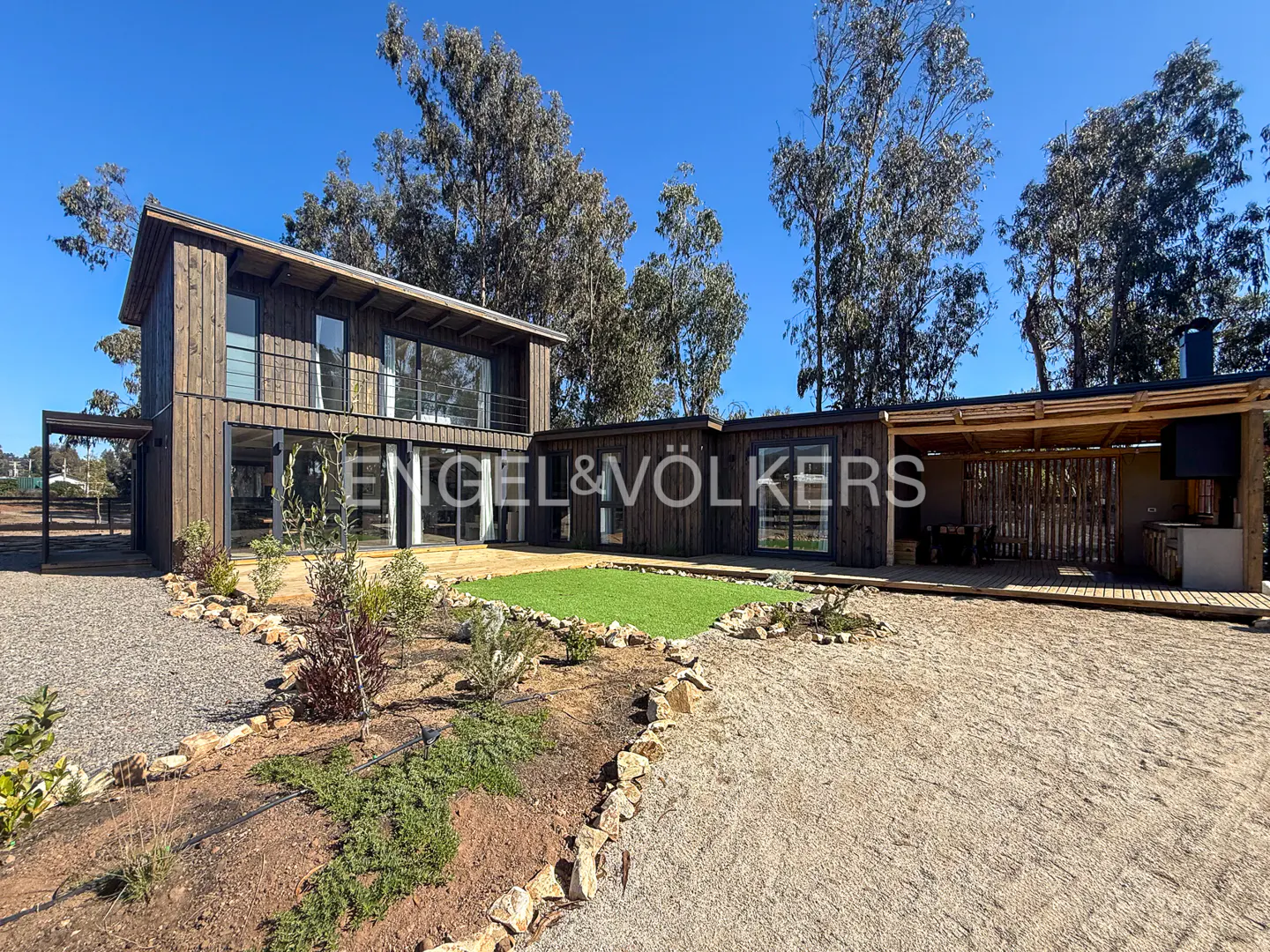 Two-story modern home with dark wood siding, large windows, and a balcony. The yard has gravel, grass, and landscaping. Blue sky and trees in the background.