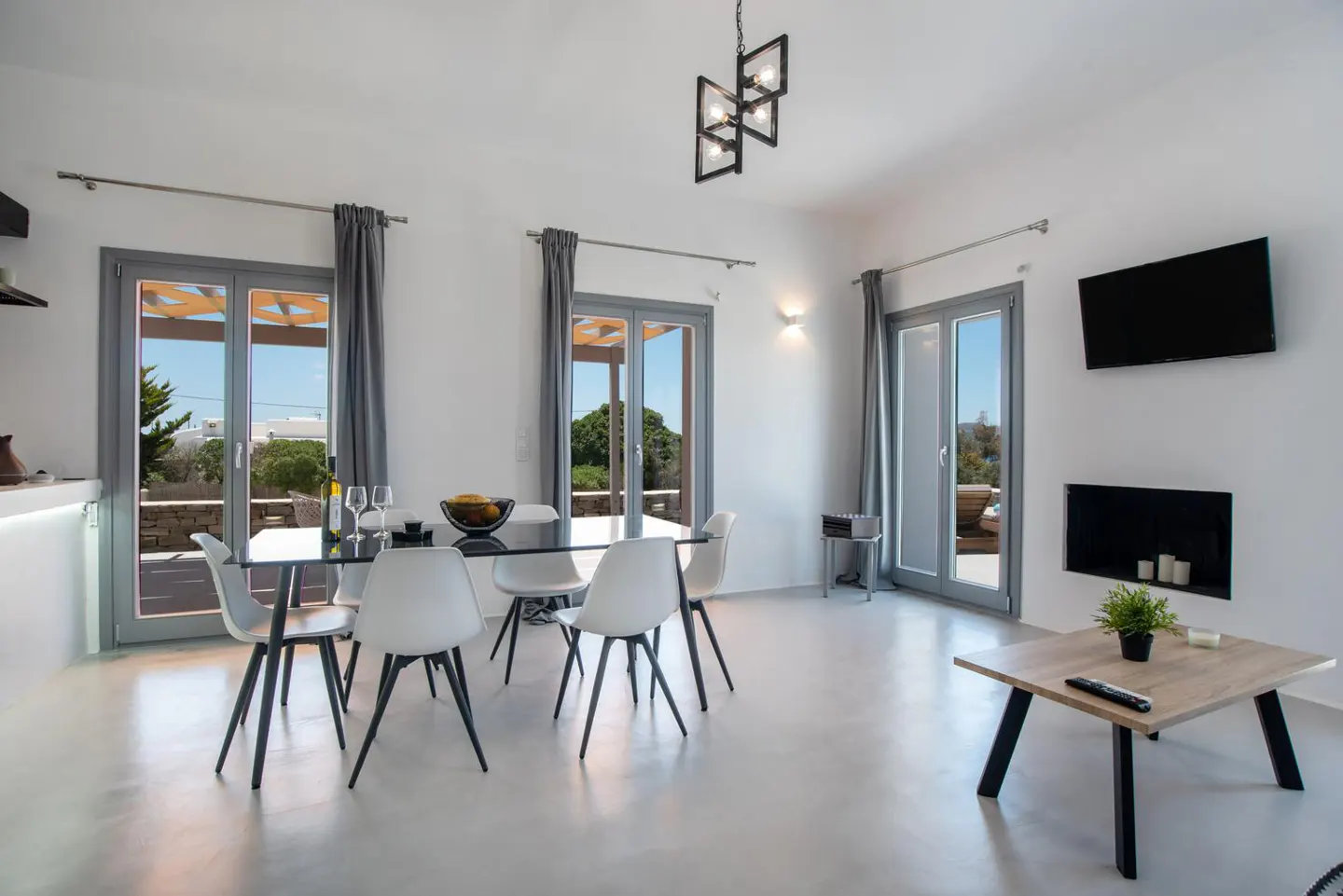Bright living room with white walls, gray trim, and polished floors. A glass table with white chairs sits near patio doors. A TV and fireplace are on the wall.