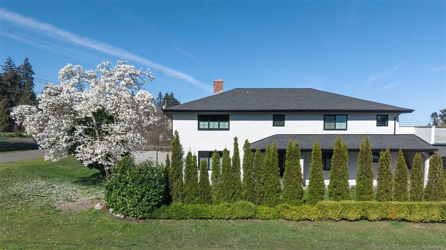 Two-story white house with a dark roof, green lawn, and a large flowering tree on a sunny day.