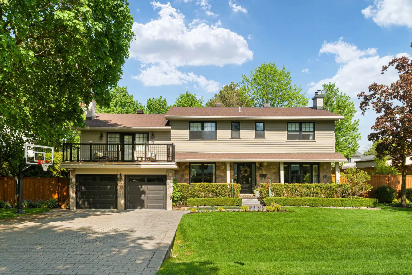 Two-story beige house with a brown roof, stone accents, and a green lawn on a sunny day. A basketball hoop is on the left.