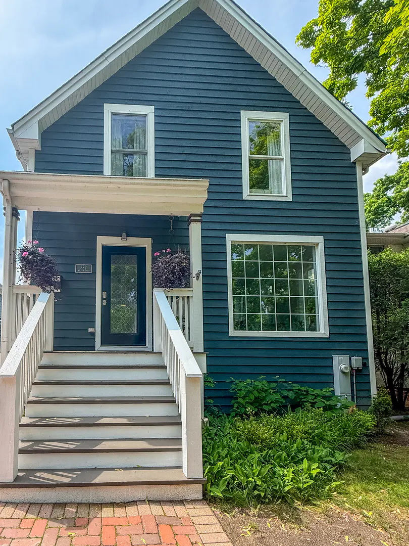 A two-story blue house with white trim, a front porch with stairs, and a brick walkway surrounded by green landscaping.