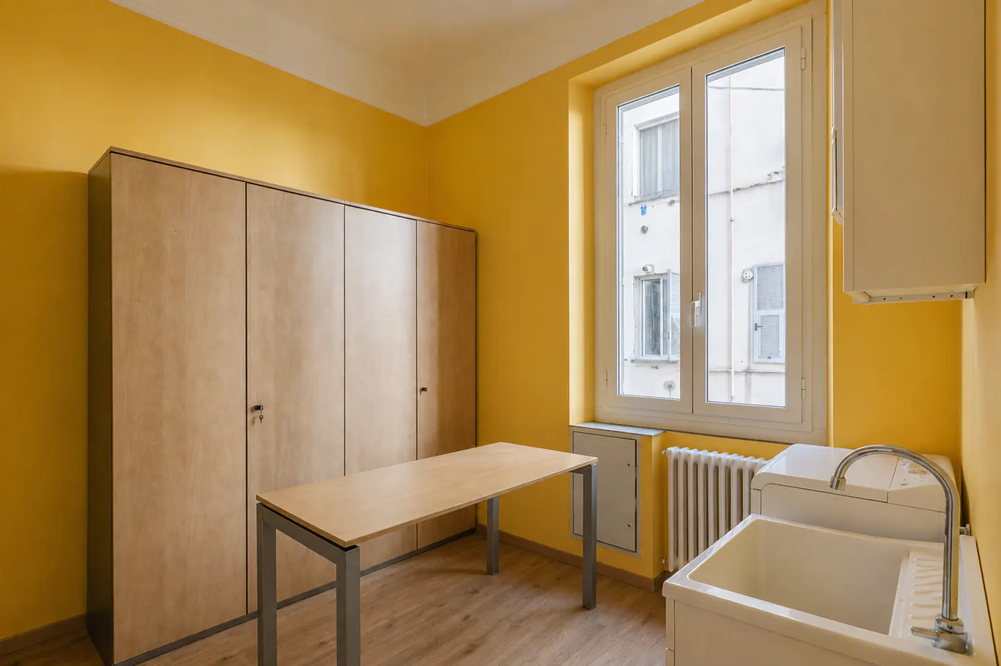 Bright yellow room with wood floors, a large cabinet, a table, a sink, and a window showing a building exterior.
